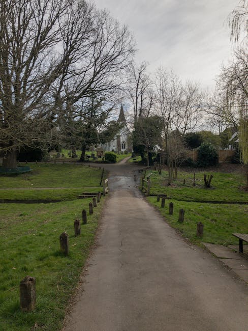A paved pathway in a park-like setting with grass and bare trees on either side, leading towards a small church with a pointed steeple in the distance. The area is surrounded by wooden posts along the